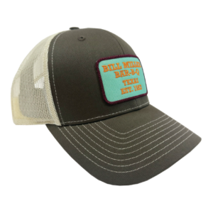 Image includes a dark tan trucker hat with a teal patch outlined with purple stitching. The patch reads, “Bill Miller Bar-B-Q Texas Est. 1953” in orange stitching.